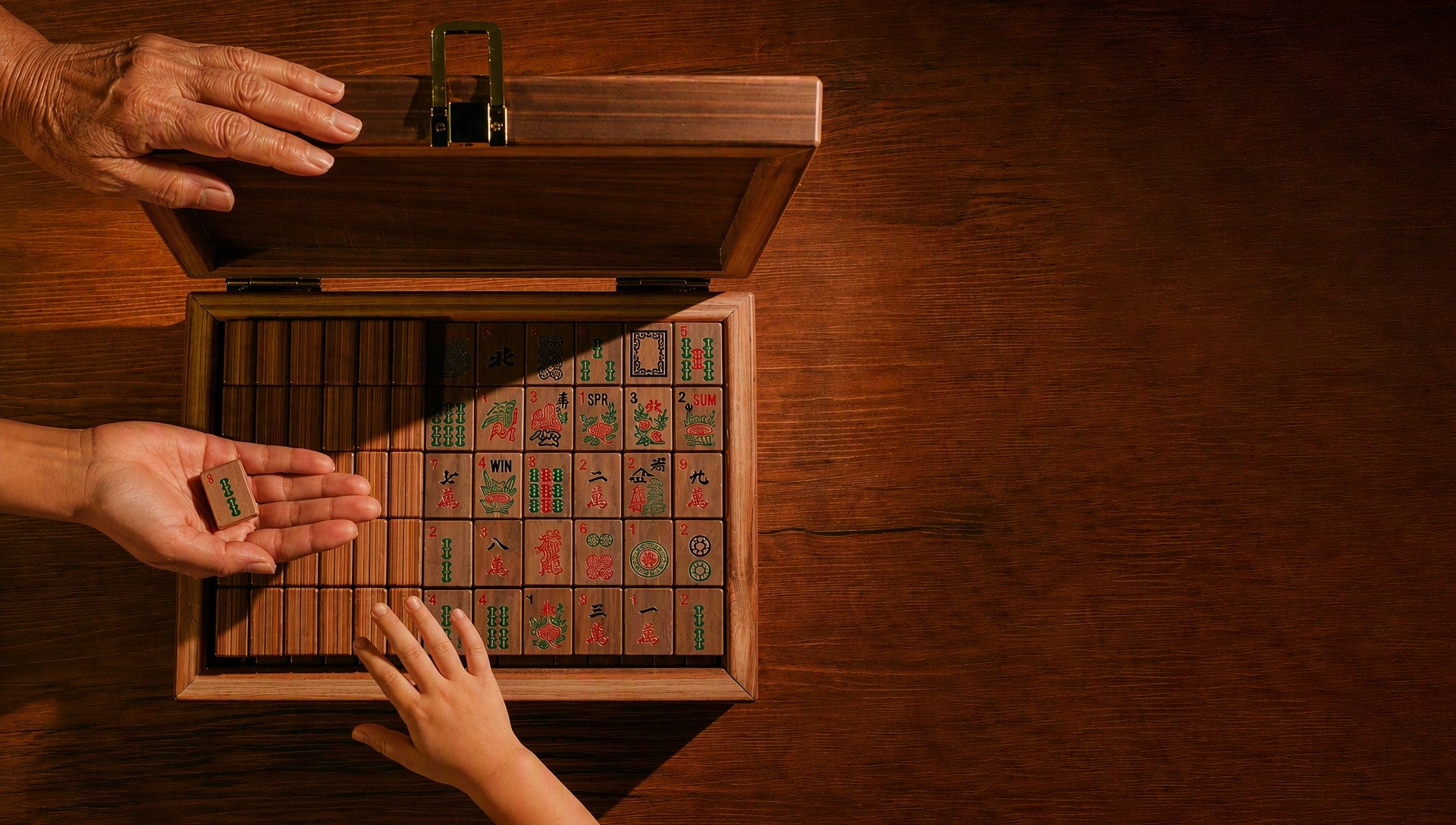 An older hand and a young child's hand lifting mahjong tiles from an open TeakTouch case on a wood-grain table, in low golden light